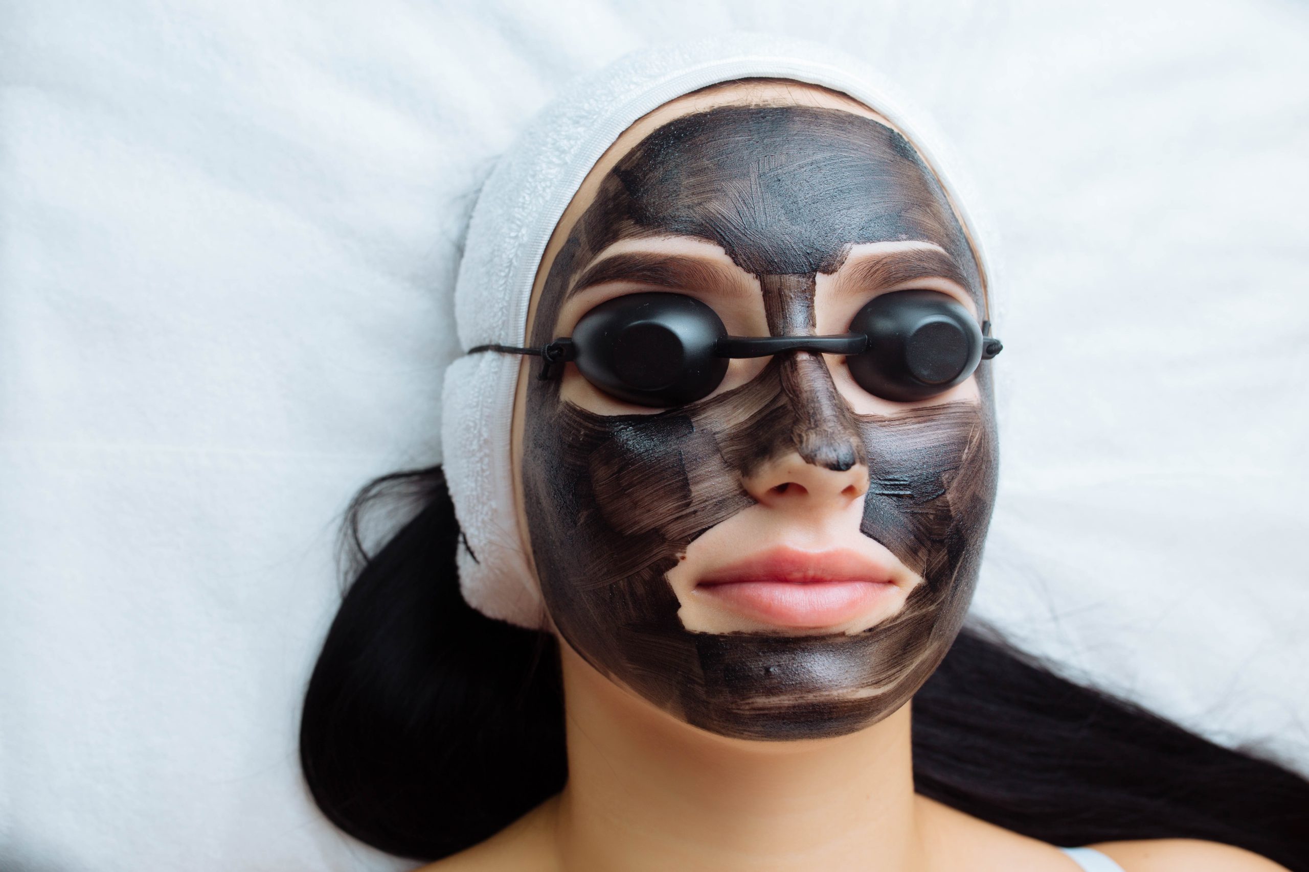 cosmetologist applying black mask on pretty woman face wearing black gloves, gorgeous woman in spa having facial procedures